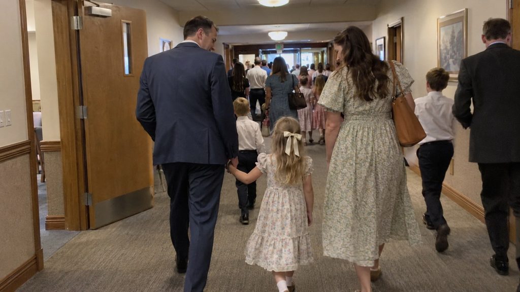 Latter-day Saint parents walking with their young daughter through a meetinghouse hallway after church.