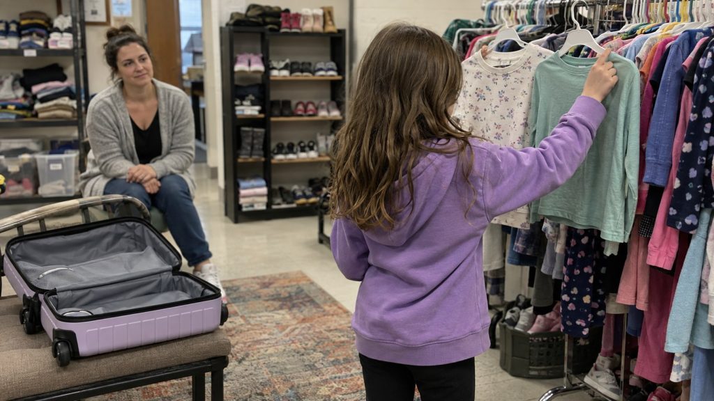 Child in a foster-care resource center choosing clothes from a rack while a volunteer waits nearby and an open suitcase sits on a bench.